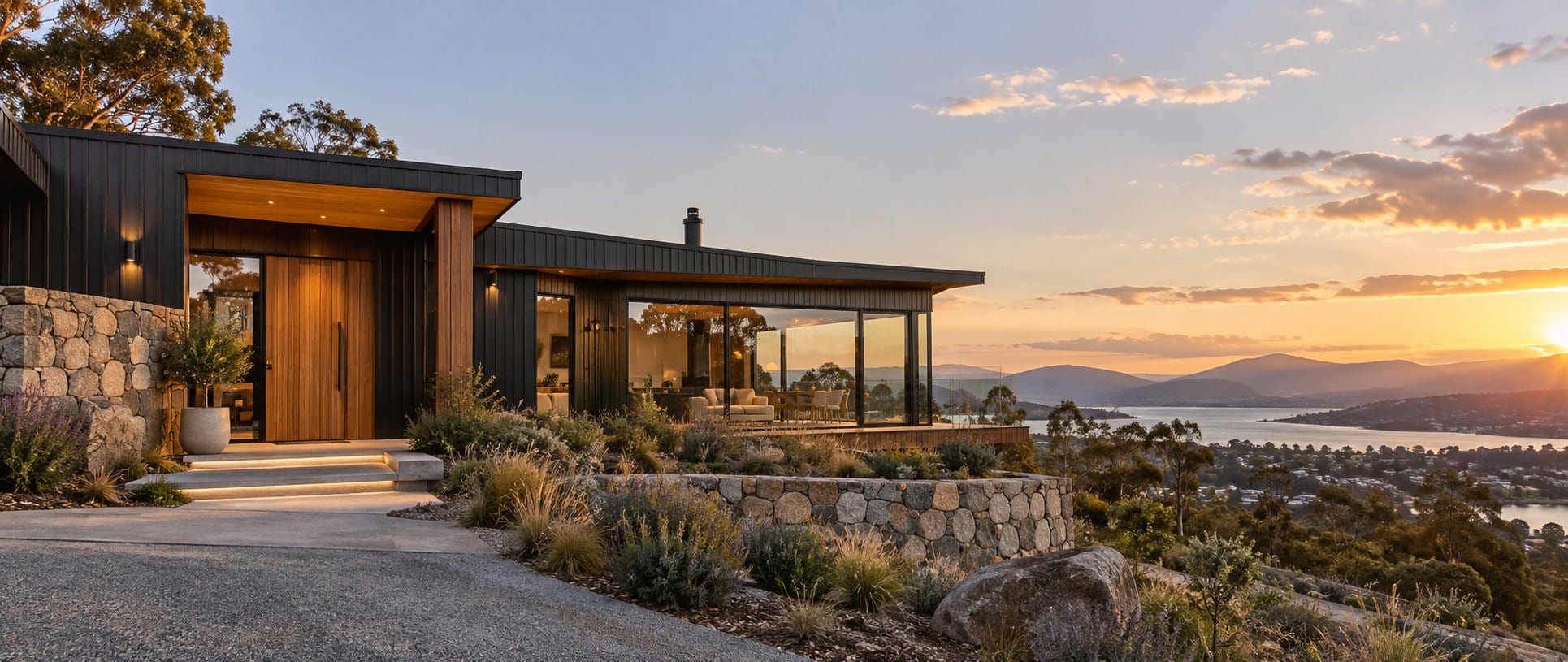 A modern dark-clad home with a timber door and stone wall at sunset, overlooking the Derwent and the hills beyond